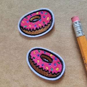 Peut inclure: Deux écussons brodés en forme de donut avec un glaçage rose et des vermicelles colorés. Les écussons ont une bordure marron et un contour blanc. Un crayon jaune avec une gomme rose est en arrière-plan.