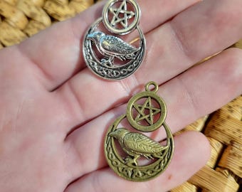 May include: Two metal pendants, one silver-toned and one bronze-toned, each featuring a circular frame with a raven perched on a crescent moon above a pentagram symbol. Both charms include a small loop at the top for attachment to a necklace.