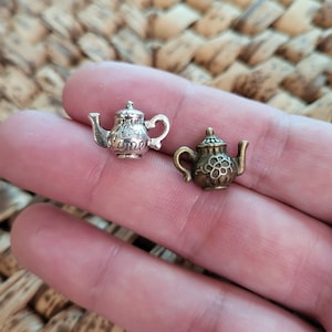 Può includere: Due ciondoli a forma di teiera in miniatura, uno argentato con le parole "Tea Time" e l'altro in bronzo con un motivo floreale.