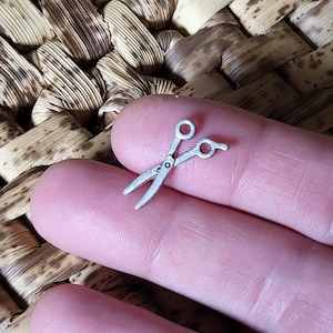 May include: A small silver-coloured pair of scissors charm. The scissors are closed and the charm is being held between two fingers.