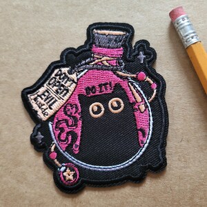 May include: Embroidered patch depicting a black cat in a pink potion bottle. The patch reads "DO IT!" and "DON'T OPEN EVIL". The design is outlined in black and has a whimsical, cartoonish style, suitable for clothing or accessories.