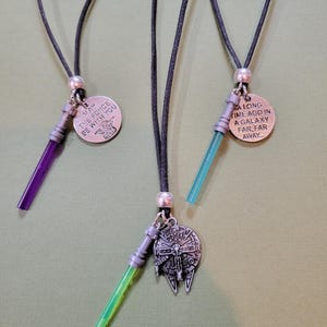 May include: Three necklaces on black cords, each featuring a silver-toned lightsaber charm with a coloured blade—purple, green, or light blue. Two necklaces include round metal tags with engraved text, while one features a spaceship charm.