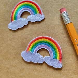 Può includere: Due toppe arcobaleno ricamate con archi rossi, arancioni, gialli, verdi, blu e viola. Ogni arcobaleno ha accenti di nuvole bianche. Una matita gialla con una gomma rosa è sullo sfondo.
