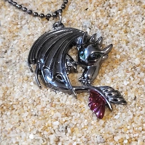 Puede incluir: Un collar de metal negro con un colgante de un dragón negro con ojos verdes y un ala roja. El dragón está enroscado con su cola envuelta alrededor de su cuerpo.