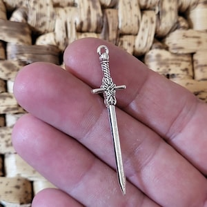 Könnte beinhalten: Ein kleiner, silberfarbener Schwertanhänger mit detailliertem Griff und spitzer Klinge. Der Anhänger hat eine Schlaufe zum Befestigen an einer Halskette oder einem Armband. Das Schwert wird in einer Hand gehalten.