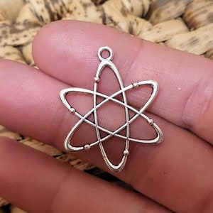 May include: A silver atom-shaped charm with orbiting rings and small spheres at the intersections. The charm is held in a person's hand against a textured background.