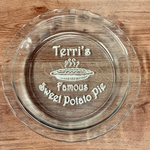 May include: A clear glass pie plate with the text "Terri's Famous Sweet Potato Pie" etched on the surface. The pie plate has a raised rim and a smooth, shiny finish.