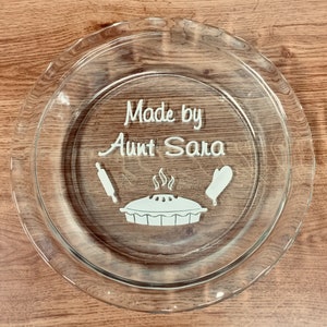 May include: A clear glass pie plate with a white design that says "Made by Aunt Sara" with a rolling pin, a pie, and an oven mitt.