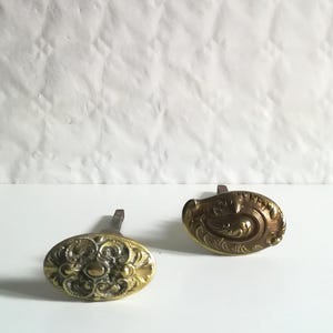 Op de afbeelding: Twee ovale antieke messing deurknoppen met sierlijke bloemmotieven. De knoppen hebben een dof gouden afwerking en staan tegen een witte achtergrond. Elke knop is ongeveer 5 cm lang.