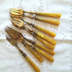 Peut inclure: Ensemble de fourchettes anciennes avec des dents dorées et des manches marbrés jaunes et ambrés. Les manches présentent des détails métalliques décoratifs. Les fourchettes sont disposées sur une nappe blanche brodée.