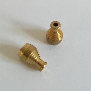 Può includere: Due piccoli oggetti metallici color ottone. Uno è in posizione verticale, l'altro è di lato. Entrambi hanno una base svasata, un corpo scanalato e una piccola apertura nella parte superiore. Gli oggetti sembrano essere componenti o parti.