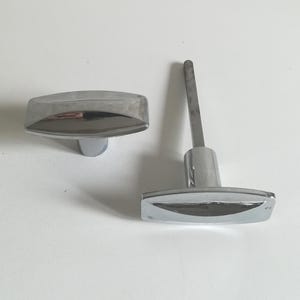 Peut inclure: Deux boutons de meuble chromés. L'un est une pièce autonome, l'autre est fixé à une tige métallique. Les deux ont un design rectangulaire et incurvé. Les boutons sont réfléchissants, suggérant une surface polie.