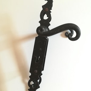 Peut inclure: Poignée de porte en fer forgé noir ornée d'une serrure et de volutes décoratives. La poignée présente un levier incurvé et des détails complexes, suggérant un style vintage ou antique. La quincaillerie est montée sur une surface blanche.
