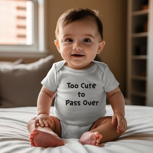 May include: A baby in a light grey onesie with the words "Too Cute to Pass Over" printed in black. The baby is sitting on a white bed, smiling. The background is blurred, showing a window and a pillow.
