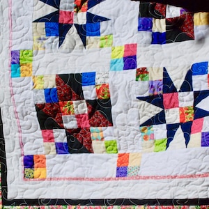 May include: A white quilt with a patchwork design featuring a variety of colorful fabrics. The quilt is quilted with a swirling pattern and has a black and white border.