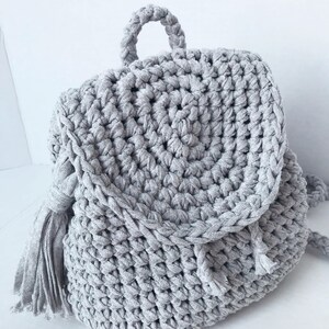 May include: A gray crocheted backpack with a round top and a single tassel on each side.