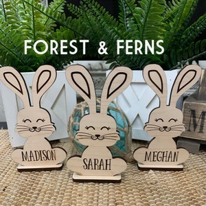 May include: Three wooden bunny figurines with personalised names, "Madison", "Sarah", and "Meghan", standing on a woven surface. The bunnies have brown ears and a smiling face.