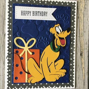 May include: A handmade birthday card featuring a cartoon dog with a yellow and orange gift box. The card has a blue background with paw prints and a banner that says "Happy Birthday".