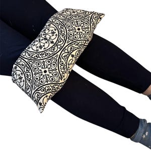May include: A rectangular, cream-colored heating pad with a black, ornate, circular pattern. The pad is resting on a person's legs, which are covered in black leggings. The background is white.