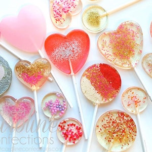 May include: A collection of colorful lollipops in various shapes, including hearts and circles. The lollipops are clear with glitter and sprinkles, in shades of pink, red, and gold. The lollipops are on white sticks, perfect for Valentine's Day or a sweet treat.