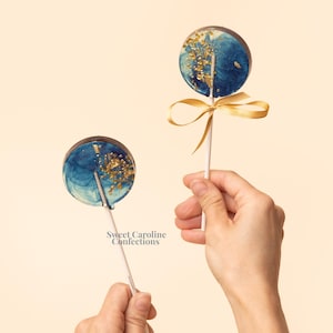 May include: Two round lollipops with blue and gold designs on white sticks. One lollipop is tied with a gold ribbon. The lollipops have a swirl of blue and gold flecks. The text "Sweet Caroline Confections" is visible.