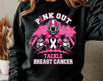 Puede incluir: Sudadera negra con un gráfico rosa. El gráfico presenta un jugador de fútbol americano con casco y protecciones, una cinta rosa y las palabras "PINK OUT" y "TACKLE BREAST CANCER". El diseño está sobre un fondo rosa salpicado.