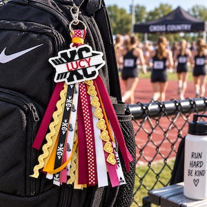 May include: A backpack charm featuring a white acrylic tag with the text LUCY and XC in black, attached to a silver lobster clasp. The charm includes multiple hanging ribbons in maroon, gold, white, and black, with patterns including hearts.
