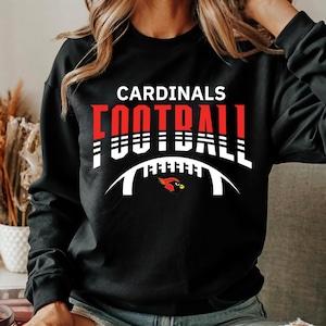 Peut inclure: Sweat-shirt noir avec un texte blanc et rouge qui dit "CARDINALS FOOTBALL" avec un contour de ballon de football blanc et un graphique de cardinal rouge.