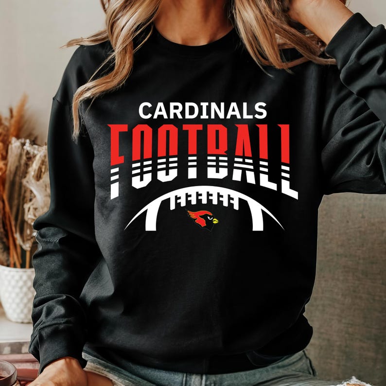Peut inclure: Sweat-shirt noir avec un texte blanc et rouge qui dit "CARDINALS FOOTBALL" avec un contour de ballon de football blanc et un graphique de cardinal rouge.