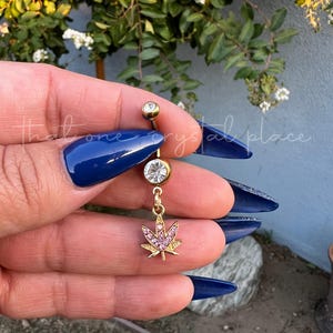 May include: Gold body jewelry with a clear gem and a dangling pink jeweled marijuana leaf charm.