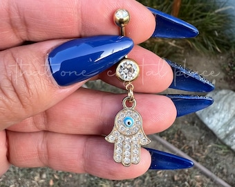 May include: Gold-toned belly button ring featuring a Hamsa hand charm. The charm is encrusted with clear crystals and a blue eye detail. The ring has a gold ball at the top and a clear crystal below.