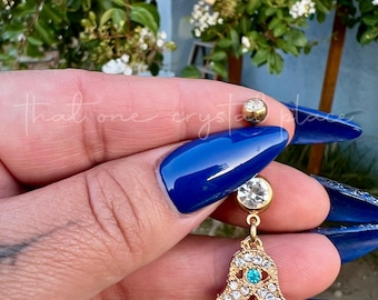 May include: Gold belly button ring with a hamsa hand charm adorned with clear and turquoise rhinestones.