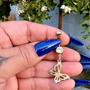 May include: Gold-colored belly button ring featuring a butterfly charm. The ring has a clear, round gem at the top and bottom, with a dangling butterfly pendant in the center. The butterfly has an openwork design.