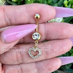 May include: Gold-colored belly button ring with a dangling heart charm. The ring features a large, clear crystal at the center and a smaller crystal at the top. The heart charm is outlined with small, clear crystals.