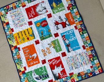 Könnte beinhalten: Eine Patchwork-Decke mit verschiedenen Dr. Seuss-Buchcovern, darunter The Cat in the Hat, The Lorax und Green Eggs and Ham. Die Decke hat einen weißen Hintergrund mit einem Rand aus bunten Buchstaben des Alphabets und einer dunkelblauen Einfassung.