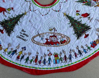 Könnte beinhalten: Ein weißer, gesteppter Weihnachtsbaumteppich mit einem bunten Aufdruck von How the Grinch Stole Christmas. Das Design zeigt grüne Grinch-Figuren, geschmückte Tannenbäume, einen roten Zug auf Schienen und eine Reihe von Whos, die Händchen halten.
