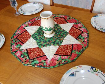 Könnte beinhalten: Eine gesteppte achteckige Tischmatte mit einem zentralen Sternmuster aus weihnachtlichem Stoff in Creme, Rot und Grün mit Stechpalmen- und Weihnachtsstern-Drucken. Eine cremefarbene Keramikvase mit Stechpalmenblatt-Design steht in der Mitte.