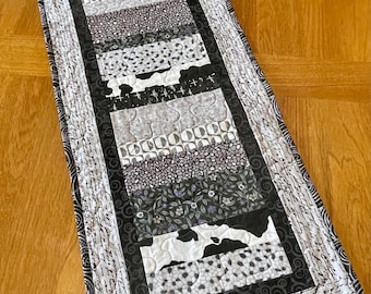 Könnte beinhalten: Ein rechteckiger, gesteppter Tischläufer mit einem Patchwork-Design in Schwarz-, Weiß- und Grautönen. Der Läufer weist verschiedene Muster auf, darunter Blumen-, geometrische und Kuhmuster, mit einem Rand aus Schrift. Der Läufer liegt auf einer Holzoberfläche.