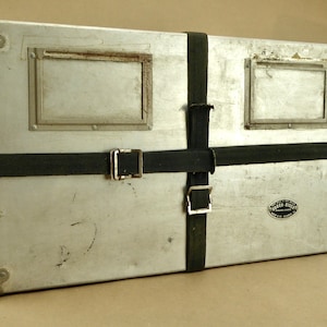 May include: A silver metal case with two rectangular windows and a black label that reads "Western Round" is secured with black straps and buckles.