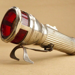 Könnte beinhalten: Eine silberne Taschenlampe mit einer roten Glasscheibe und einer Metallhalterung. Die Taschenlampe hat einen gerippten Körper und einen kleinen Schalter an der Seite.