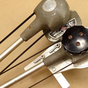 May include: Two vintage drafting machines, likely used for technical drawing. The machines have a grey and black colour scheme with silver metal arms and black belts. One machine has a circular dial with degree markings.