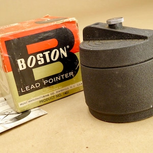 Puede incluir: Sacapuntas Boston Lead Pointer vintage con caja original. La caja es roja, negra y crema con el texto "BOSTON LEAD POINTER". El sacapuntas es negro con un botón plateado.