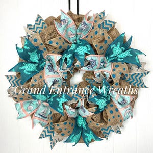 May include: A decorative wreath featuring natural burlap mesh with teal polka dots and chevron patterns. The wreath is accented with wired ribbons displaying sea turtles, anchors, sailboats, and ship wheels. The text Grand Entrance Wreaths is visible.