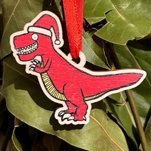 May include: A wooden Christmas ornament depicting a red Tyrannosaurus Rex wearing a Santa hat. The dinosaur has a white outline and a yellow striped belly. A red ribbon is attached for hanging. The ornament is set against a background of green leaves.