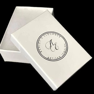 May include: A white jewelry box with the lid slightly ajar, revealing the interior. The lid features a circular logo with the text "Ashley's Elegance Artisan Jewelry" in black. The box is set against a black background.