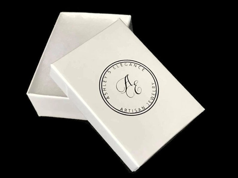 May include: A white jewelry box with the lid slightly ajar, revealing the interior. The lid features a circular logo with the text "Ashley's Elegance Artisan Jewelry" in black. The box is set against a black background.