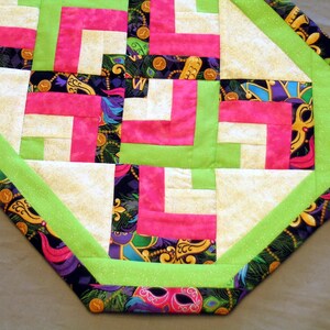 May include: A green, pink, and white patchwork table runner with a Mardi Gras themed fabric border. The border features gold, purple, and green colors with a repeating pattern of masks, feathers, and beads.