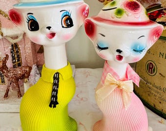 May include: Two vintage ceramic cat figurines. One cat is wearing a yellow sweater with a black tassel, and the other is wearing a pink sweater with a pink bow. Both cats have white faces, blue eyes, and hats decorated with flowers.