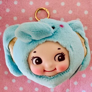 Może przedstawiać: Jasnoniebieski pluszowy brelok do kluczy z uśmiechniętą twarzą lalki w kapturze z uszami słonia. Lalka ma r&oacute;żowe policzki, duże oczy i małe czerwone usta. U g&oacute;ry przymocowany jest złoty pierścień.