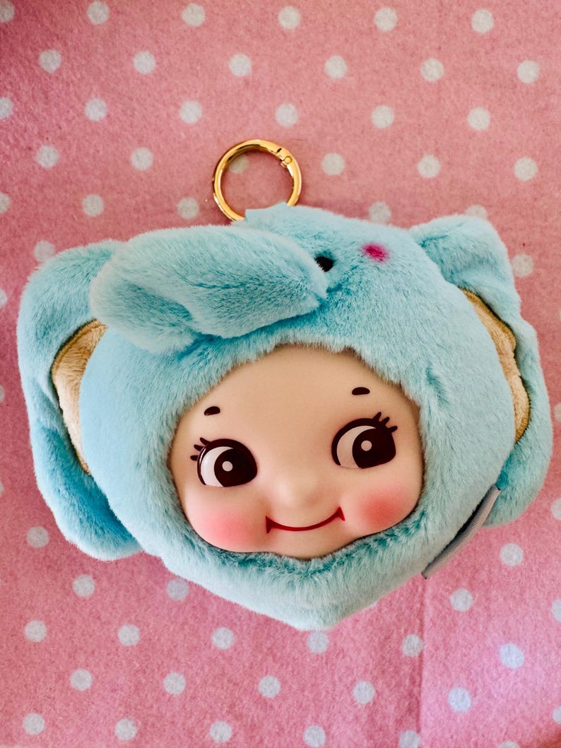 Może przedstawiać: Jasnoniebieski pluszowy brelok do kluczy z uśmiechniętą twarzą lalki w kapturze z uszami słonia. Lalka ma r&oacute;żowe policzki, duże oczy i małe czerwone usta. U g&oacute;ry przymocowany jest złoty pierścień.